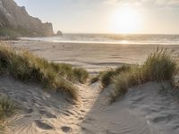 a sandy beach with a sun setting over the water and sand dunes, grass, and mountains