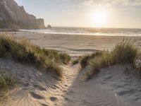 a sandy beach with a sun setting over the water and sand dunes, grass, and mountains