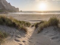 a sandy beach with a sun setting over the water and sand dunes, grass, and mountains