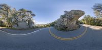 an asphalt road that has a rock in the middle with trees in the background, in front of some rocks and a sky