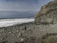 two people walking near the ocean by the rocky coastline and the cliffs in front are covered with large, blue rocks