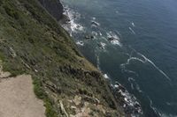 a man walking along a path beside the ocean in a scenic landscape area with a steep cliff to one side