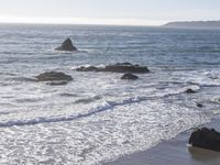 some rocks are sitting on the beach near the water's edge as a surfer holds a surfboard