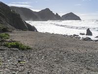 a rock cliff is over the water and has a sandy beach next to it with green vegetation, rocks and sea