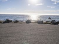 a person sitting on the curb near the ocean with a surfboard behind them, as the sun shines and rocks look like rocks