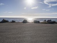 a person sitting on the curb near the ocean with a surfboard behind them, as the sun shines and rocks look like rocks