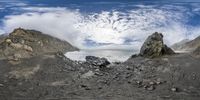 three mountain ranges with sea, rocks and snow on them under a blue sky filled with clouds