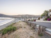 a wooden fence and sidewalk next to the beach and ocean in front of houses at sunset