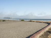 a sidewalk with a curb that runs alongside the beach and ocean, which is partly wet, the sun shines