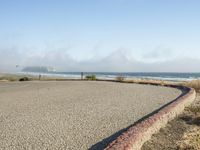 a sidewalk with a curb that runs alongside the beach and ocean, which is partly wet, the sun shines