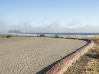 a sidewalk with a curb that runs alongside the beach and ocean, which is partly wet, the sun shines