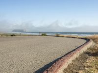 a sidewalk with a curb that runs alongside the beach and ocean, which is partly wet, the sun shines