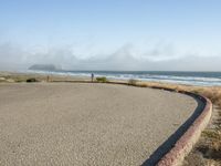 a sidewalk with a curb that runs alongside the beach and ocean, which is partly wet, the sun shines