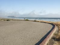 a sidewalk with a curb that runs alongside the beach and ocean, which is partly wet, the sun shines