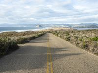 an empty road on the side of the beach and hills in the distance, with green grass in front of the road