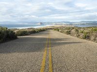 an empty road on the side of the beach and hills in the distance, with green grass in front of the road