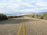 an empty road on the side of the beach and hills in the distance, with green grass in front of the road