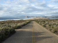 an empty road on the side of the beach and hills in the distance, with green grass in front of the road