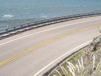 the lone yellow bus is driving on the paved road near the ocean near a metal railing