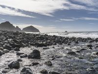 an ocean and rocks coast with some surfers in the water with blue skies in background