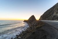 a road on the side of a cliff with a sunset in the background near the ocean