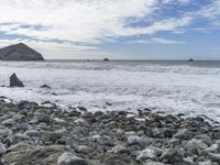 a lone surfboard is seen on the rocky beach shore, with a lighthouse in the background