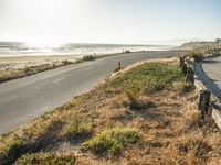 a couple of people are riding motorcycles on the road by the beach, and a long bench is sitting on the side of the road