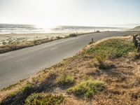 a couple of people are riding motorcycles on the road by the beach, and a long bench is sitting on the side of the road