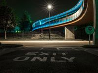 the curved bridge is lit up blue with green light on it's sides and is over a road in front of a concrete wall