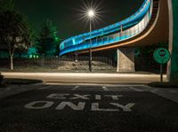 the curved bridge is lit up blue with green light on it's sides and is over a road in front of a concrete wall