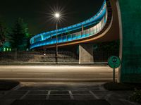 the curved bridge is lit up blue with green light on it's sides and is over a road in front of a concrete wall