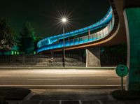 the curved bridge is lit up blue with green light on it's sides and is over a road in front of a concrete wall