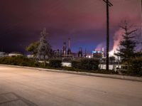 a street scene with cars, a factory and trees at night in the background with sky full of smoke