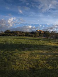 a large grassy field with green grass under blue skies in the distance is trees and hills and buildings