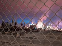 an oil refinery is seen through a chain link fence in this city photo, it appears a red and blue sky is visible in the background