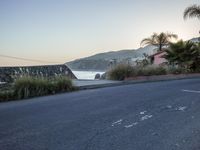 a road on the edge of a seawall, near some rocks and houses with palm trees