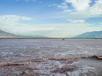 an open area with some water and some rocks near some mountains with a sky background