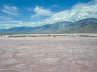 the desert in front of a large mountains is pink and gray with some clouds above