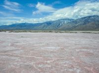 the desert in front of a large mountains is pink and gray with some clouds above