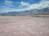 the desert in front of a large mountains is pink and gray with some clouds above