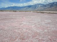 the desert in front of a large mountains is pink and gray with some clouds above