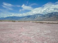 the desert in front of a large mountains is pink and gray with some clouds above