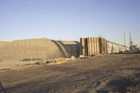 a group of pipe piles in front of a building under construction on dirt ground with metal poles nearby