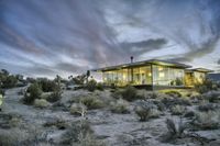 a home in the middle of nowhere surrounded by plants and trees in the desert as the sun sets