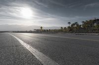 a sun reflecting off an empty roadway in the middle of desert with mountains and palm trees