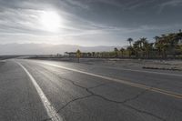 a sun reflecting off an empty roadway in the middle of desert with mountains and palm trees