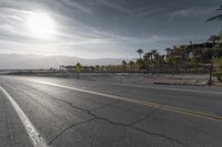 a sun reflecting off an empty roadway in the middle of desert with mountains and palm trees