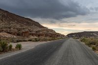 a person riding a bicycle down a deserted road between large rocks and desert plants under a cloudy sky