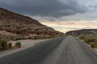 a person riding a bicycle down a deserted road between large rocks and desert plants under a cloudy sky