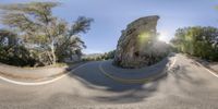 a curved road surrounded by trees with the sun going down on the cliff as a man riding a motorcycle rides on it